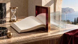 Libro contable de papel algodón crema atravesando un marco de cristal burdeos y latón junto a un reloj de arena sobre piedra travertino